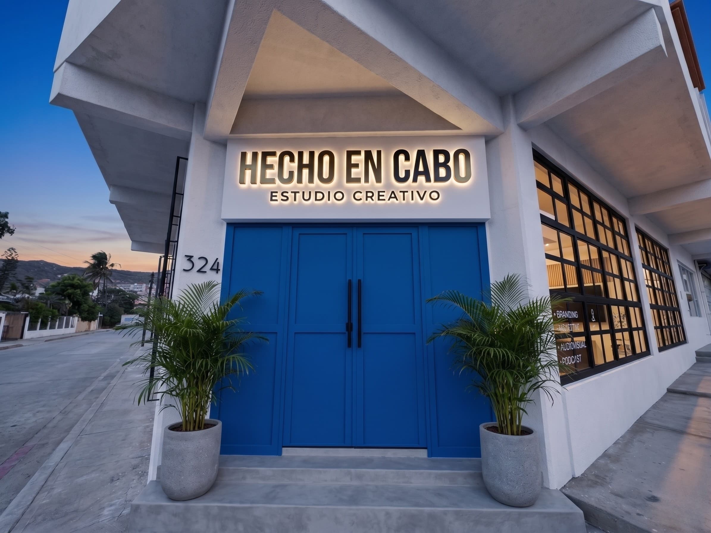 Fachada del taller de Hecho en Cabo al atardecer: portón azul, rótulo iluminado que dice Hecho en Cabo Estudio Creativo, y el número 324 sobre el muro blanco